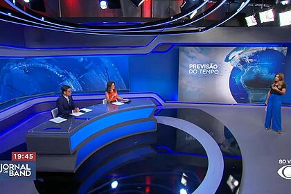 Rodolfo Schneider e Adriana Araújo sentados na bancada do Jornal da Band recebendo Joana Treptow na Previsão do Tempo