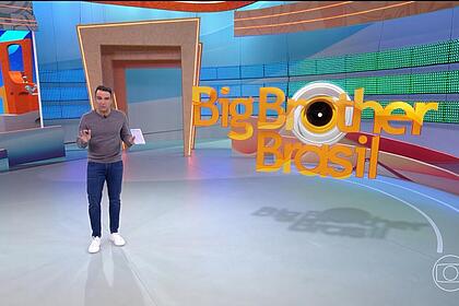 Tadeu Schmidt no centro do BBB 24, com as mãos levantadas, explicando a dinâmica da semana