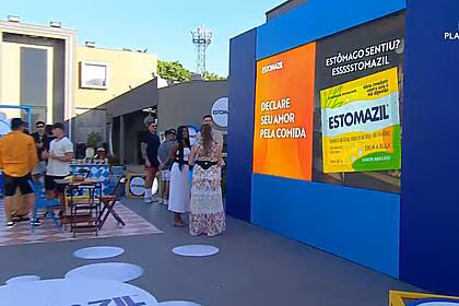 Cenário da atividade patrocinada pelo estomazil na área externa da mansão do a grande conquista contendo mesinhas, pastel, hambúrguer e muito mais