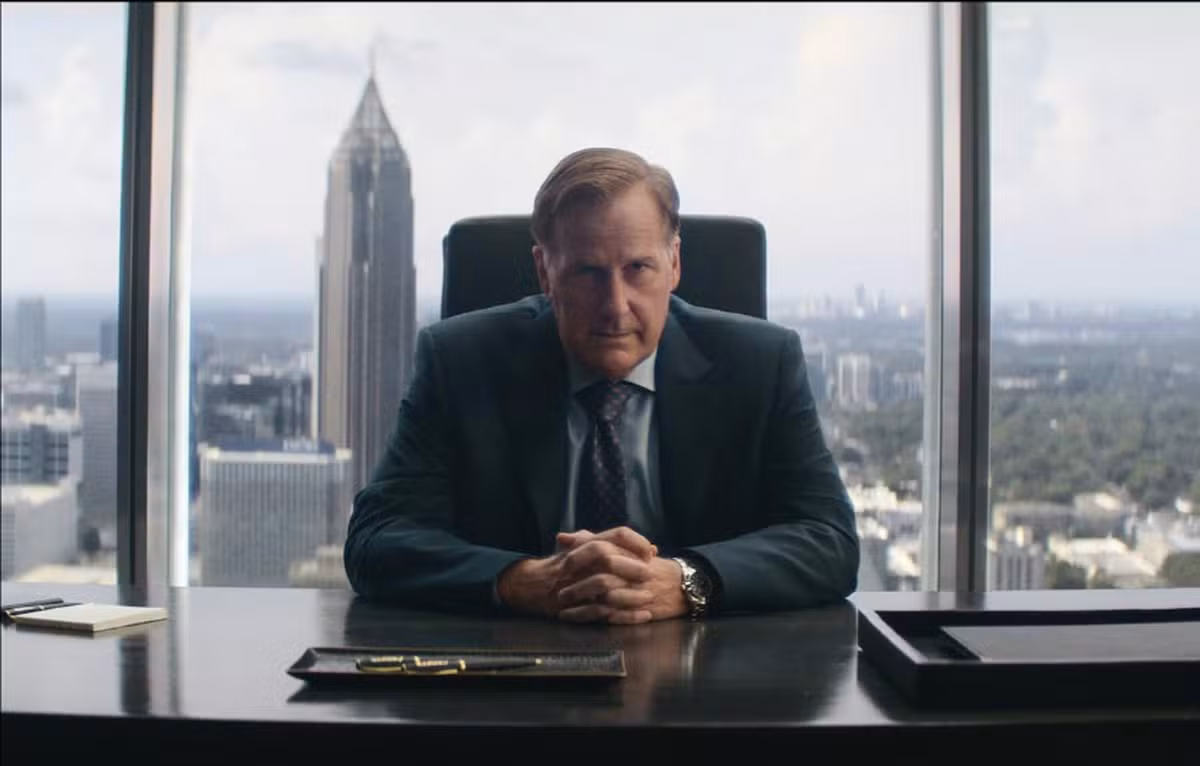 A imagem mostra Jeff Daniel de terno sentado em sua mesa de escritório. Ao fundo, vê se a cidade ao fundo de uma vista bem alta.