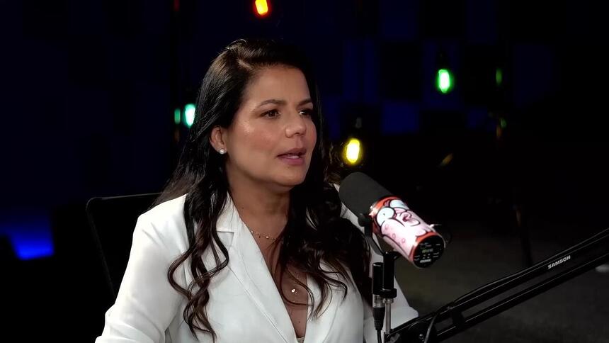 Nívea Stelmann, vestindo um blazer branco, sentada em frente a um microfone em um estúdio iluminado por luzes azuis e verdes desfocadas. A atriz está em uma sessão de gravação do podcast Inteligência Ltda., onde ela revelou sua decisão de recusar uma proposta da Playboy