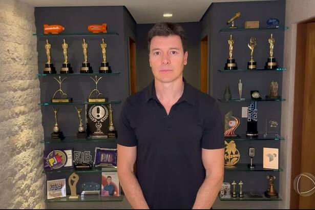 Rodrigo Faro com semblante triste em frente ao painel com troféus. Ele veste uma camisa preta em homenagem a Silvio Santos