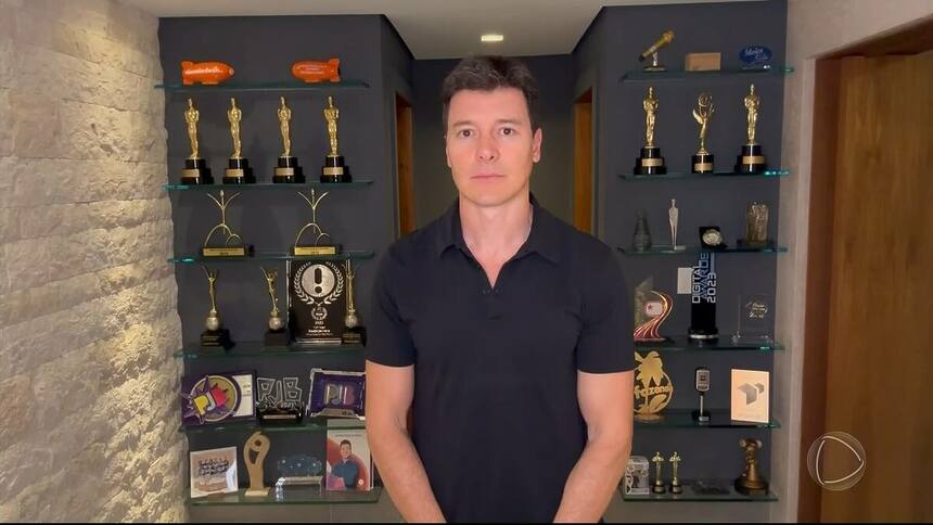 Rodrigo Faro com semblante triste em frente ao painel com troféus. Ele veste uma camisa preta em homenagem a Silvio Santos