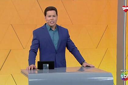 Marcão do Povo em pé, atrás da bancada no estúdio do Tá na Hora