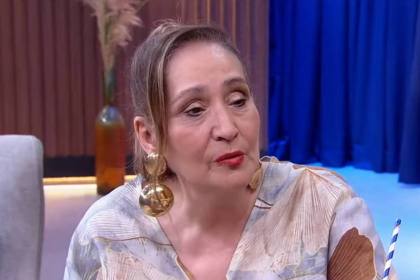 Sonia Abrão, com uma roupa em tom claro, sentada em um sofá no estúdio do Companhia Certa