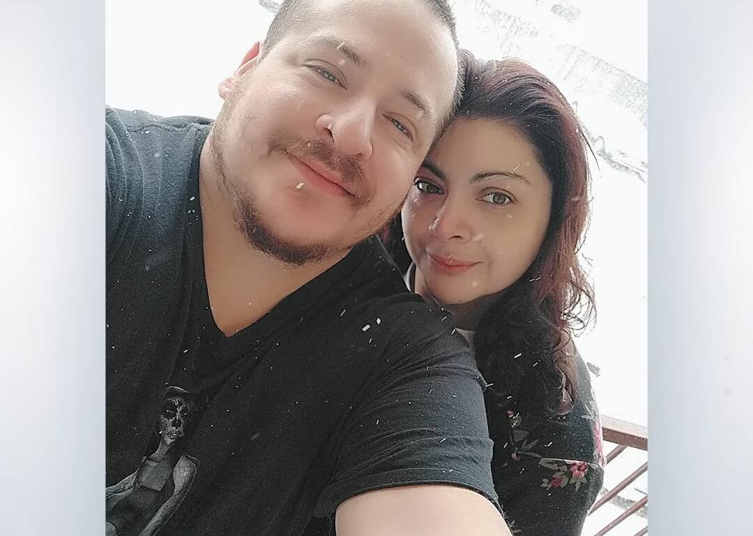 Karina ao lado de um homem fazendo selfie. Eles estão de camisa e blusa preta