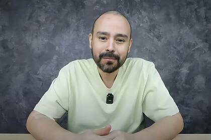 Klenion Livio de camisa bege sentado com semblante triste, em sua bancada, gravando para o YouTube