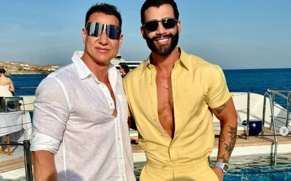 Rogerinho e Gusttavo Lima sorridentes, lado a lado, em um iate; ambos usam óculos escuros, o segurança está de camisa branca, enquanto o calor está de camisa amarela