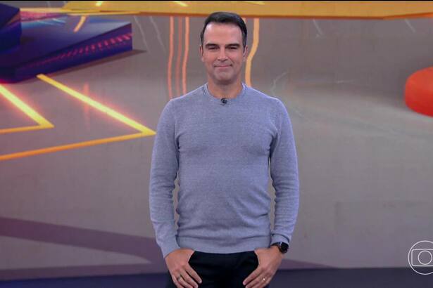 Tadeu Schmidt de camisa cinza, em pé e sorridente, no cenário do BBB 25