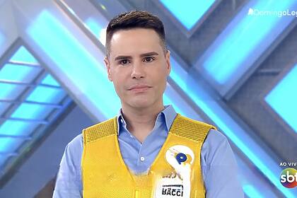 Luiz Bacci com olhar triste no palco do Domingo Legal. Ele veste camisa azul com o colete do Passa ou Repassa