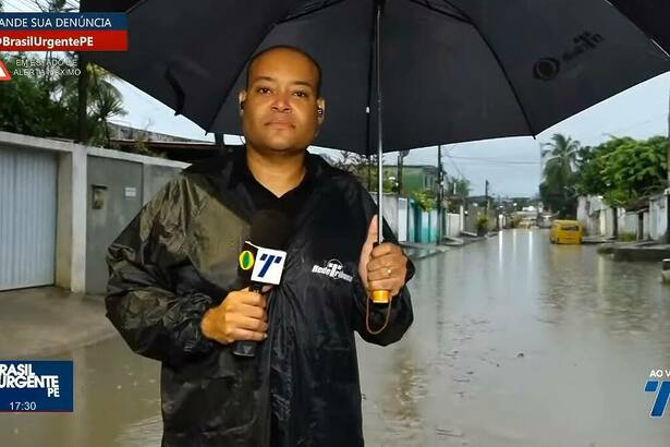 Apresentador Moab Augusto nas ruas de Recife, usando casaco de chuva e segurando um guarda-chuva e o microfone da TV Tribuna Band, apresentando o Brasil Urgente PE