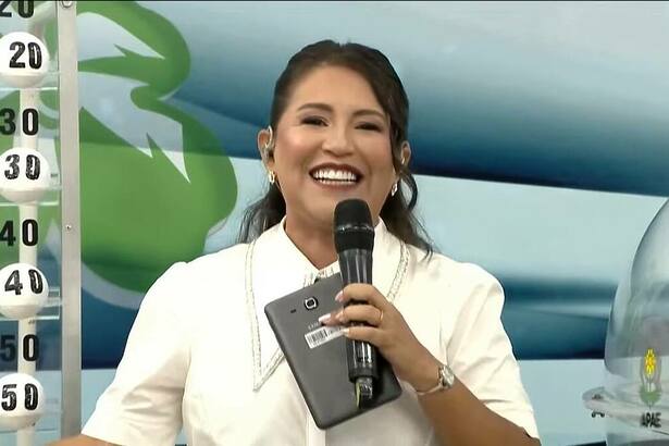 Apresentadora do Alagoas dá Sorte com um tablet e microfone nas mãos e sorrindo, antes de iniciar o sorteio