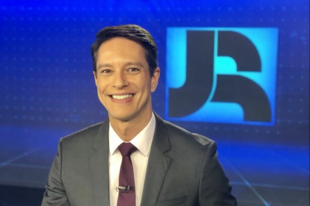Sergio Aguiar sorridente no cenário do Jornal da Record em frente ao logo do telejornal