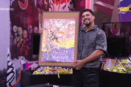 Vinicius segura quadro de fitas com desenho de Renata no quarto Barrado no Baile do BBB 25. O brother sorrir para a câmera