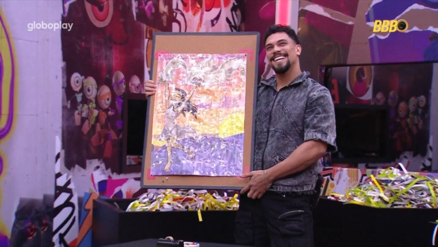 Vinicius segura quadro de fitas com desenho de Renata no quarto Barrado no Baile do BBB 25. O brother sorrir para a câmera