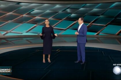 Carolina Ferraz, de vestido preto e com as mãos abertas ao lado de Roberto Cabrini na apresentação do Domingo Espetacular