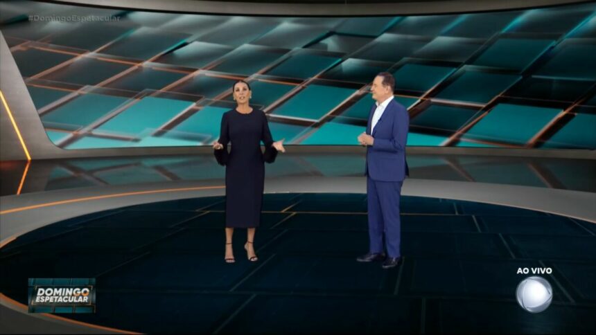 Carolina Ferraz, de vestido preto e com as mãos abertas ao lado de Roberto Cabrini na apresentação do Domingo Espetacular