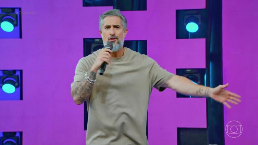 Marcos Mion com os braços abertos no palco do Caldeirão falando de cantores sertanejos