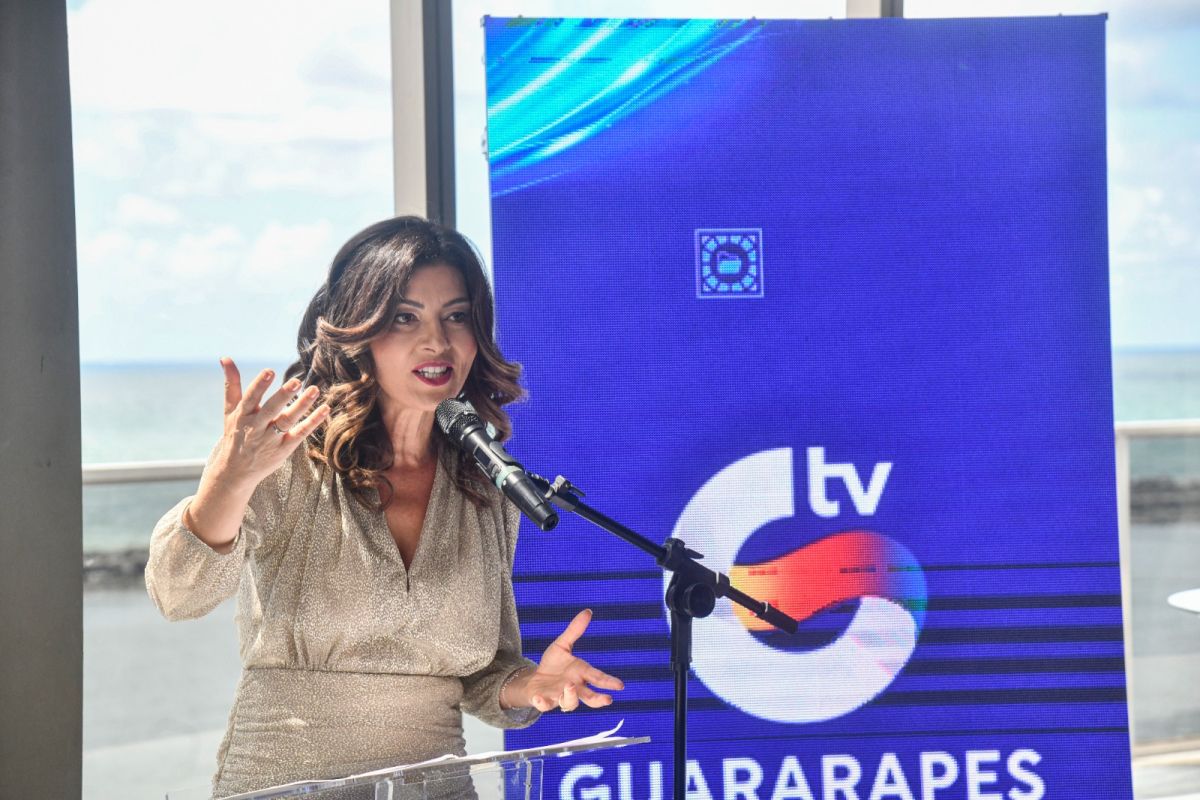 Meiry Lanunce fala ao microfone do lado do logo da TV Guararapes