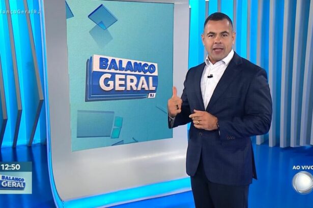 Tino Junior, de terno preto e camisa azul, fazendo sinal de legal, em frente ao telão do Balanço Geral RJ na RECORD Rio