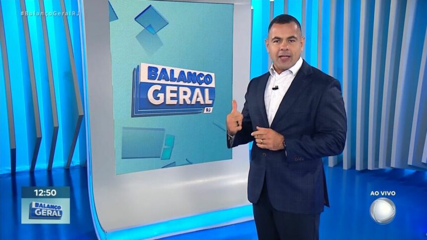 Tino Junior, de terno preto e camisa azul, fazendo sinal de legal, em frente ao telão do Balanço Geral RJ na RECORD Rio