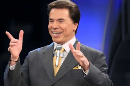 Silvio Santos com os braços levantados e o tradicional microfone colado na roupa. Ele está sorrindo