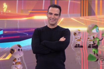 Tadeu veste camisa preta e sorrir em frente aos dummies no cenário do bbb 25