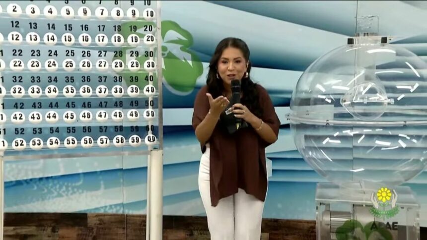 Apresentadora do Alagoas dá Sorte, de blusa marrom e sorrindo, ao lado do painel expositor com as bolas do sorteio