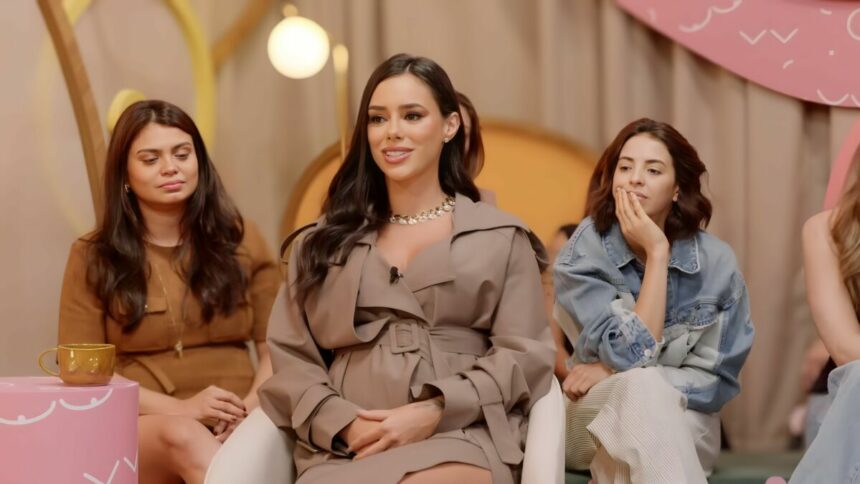 Bruna Biancardi de vestido bege, sorrindo, enquanto fala de Neymar em um podcast; há duas mulheres por trás observando Bruna