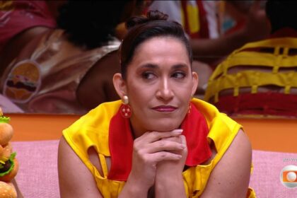 Daniele com os braços próximos a boca sentada no sofá da sala do bbb 25 respondendo perguntas de tadeu