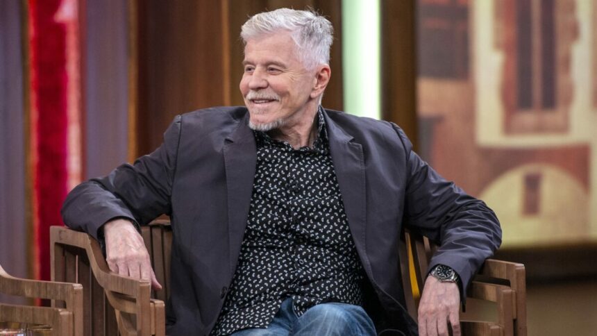 Miguel Falabella de calça jeans e blazer preto sentado em uma cadeira