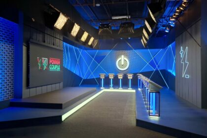 Sala de Apostas do Power Couple Brasil 7 com luzes de neon verde e azul, telão com logo do Power Couple e púlpitos onde os casais fazem apostas