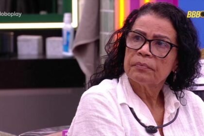 Vilma veste blusa branca e olha para tela do quarto do líder do bbb 25 com olhar de desdém, onde vinicius aparece