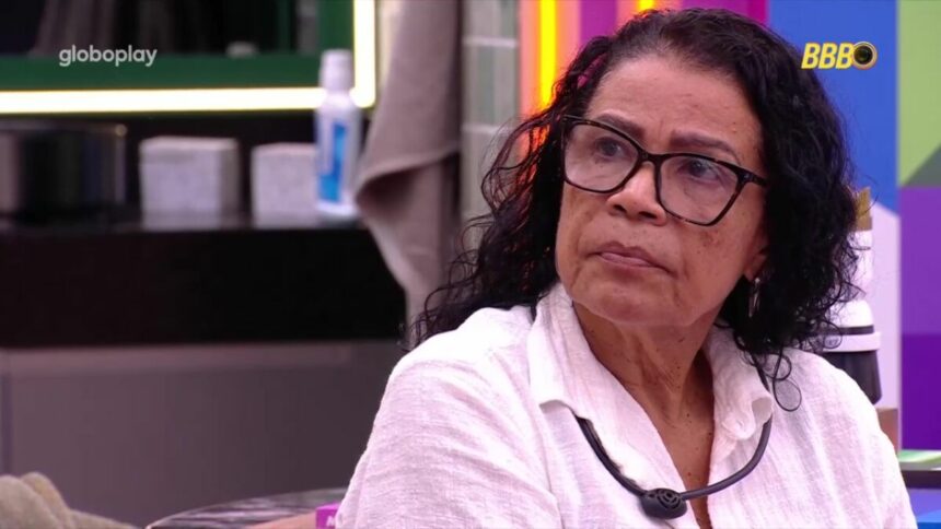 Vilma veste blusa branca e olha para tela do quarto do líder do BBB 25 com olhar de desdém, onde Vinicius aparece