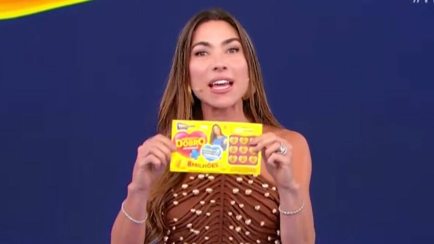 Patricia Abravanel sorrindo e segurando o bilhete do quarto sorteio da Tele Sena de Mães 2025. Ela veste um vestido marrom.