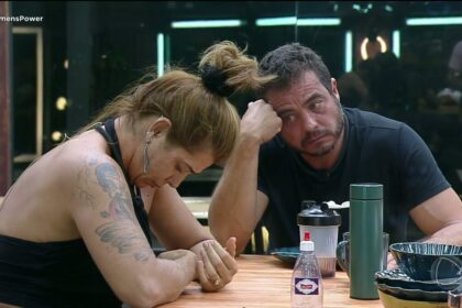 Adriana de cabeça baixa sentada na mesa de jantar do Power Couple Brasil, e Dhomini olhando para ela