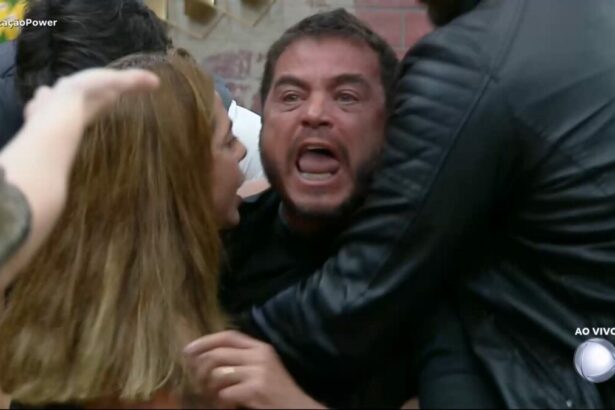 Dhomini gritando contra Carol e sendo segurado por outros participantes no sofá do Power Couple