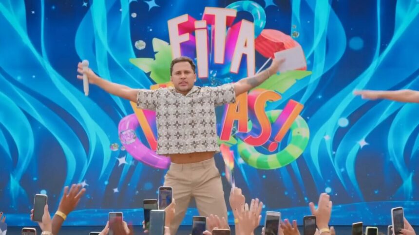 Lucas Guimarães abre os braços em frente ao telão do programa Eita,Lucas! no SBT. Ele está com semblante assustado e segura um microfone branco em uma das mãos