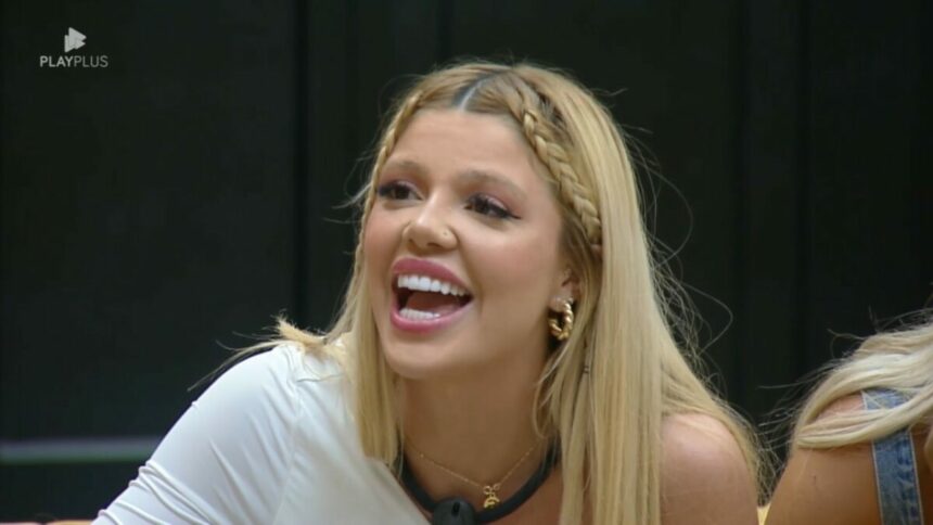 Giovanna de blusa branca, sentada no sofá da Mansão Power, sorrindo ao ver Diego que disputava a Prova dos Homens