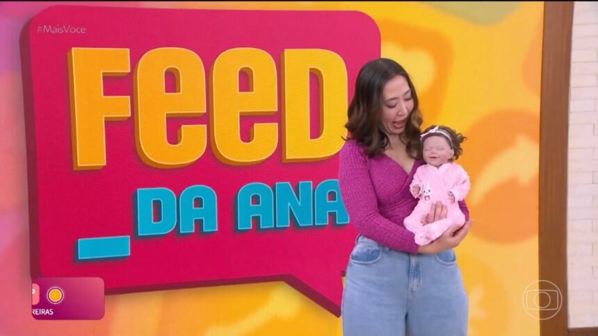 Juju Massaoka segura Bebê Reborn em frente ao telão do Feed da Ana do Mais Você. A repórter olha o bebê com a boca aberta