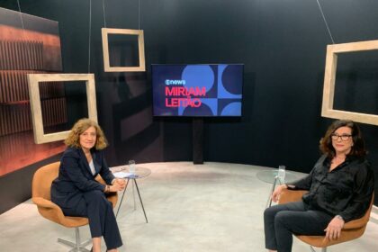 Miriam Leitão sentada em uma cadeira no cenário da Globo News ao lado da presidente da Petrobras, Magda Chambriard