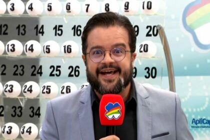 Apresentador do Pernambuco da Sorte em frente ao painel com as bolas do sorteio de 11 de maio. Ele segura um microfone vermelho