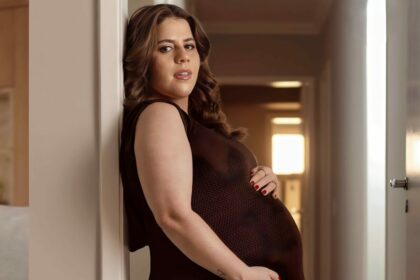 Tati Machado com a mão na barriga ainda grávida. Ela veste um vestido marrom e está encostada em uma parede