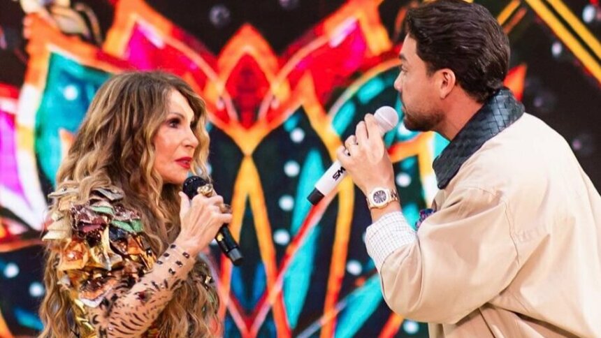 Elba Ramalho e Wesley Safadão cantando enquanto um olha para o outro. Eles usam roupas características de São João