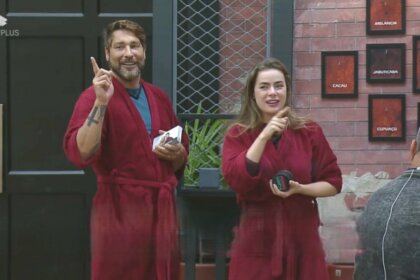 Victor e Rayanne seguram as placas dos quartos do Power Couple Brasil 7. Eles vestem roupão vermelho do casal Power e estão sorrindo
