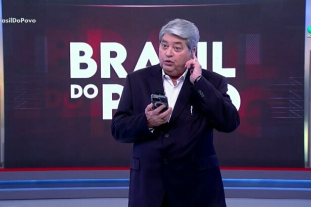 Datena coça a orelha, levemente irritado, enquanto segura um smartphone na mão no centro do cenário do Brasil do Povo na redeTV!
