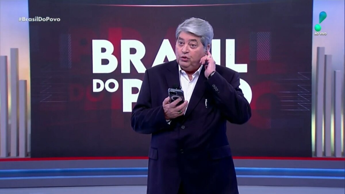 Datena coça a orelha, levemente irritado, enquanto segura um smartphone na mão no centro do cenário do Brasil do Povo na redeTV!