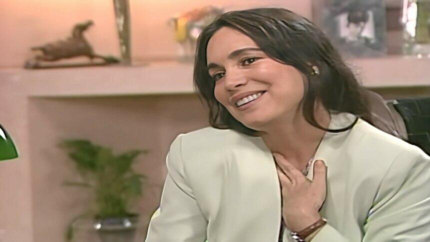 Helena, Regina Duarte, com a mão no peito em ar de felicidade. Ela está sentada no escritório de Carlos em cena de História de Amor