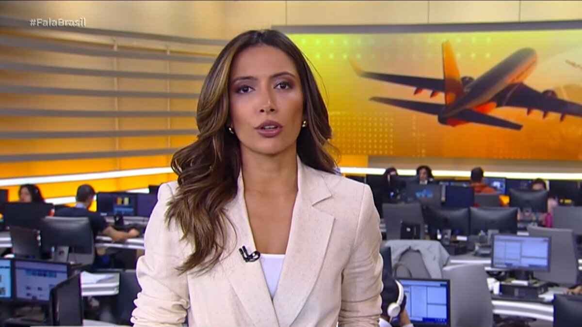 Paloma Poeta veste blazer branco no cenário do Fala Brasil. Ao fundo a redação com o telão do telejornal