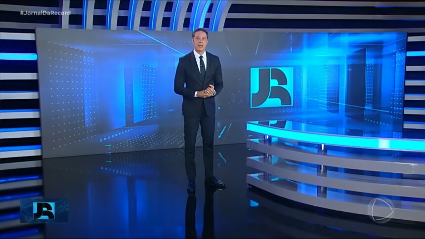 Sergio Aguiar em pé no centro do cenário do jornal da Record vestindo terno preto alguns dias antes de sua demissão. Ele está com semblante sério.